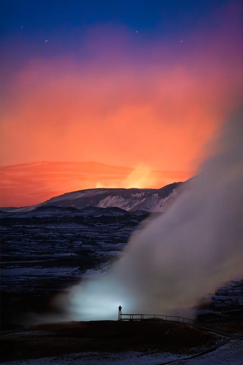 A person stands in front of a column of steam as lava shoots into the air on the horizon.