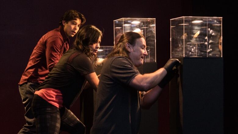 Three people, wearing black and red, appear to be trying to unlock glass cases in a black room