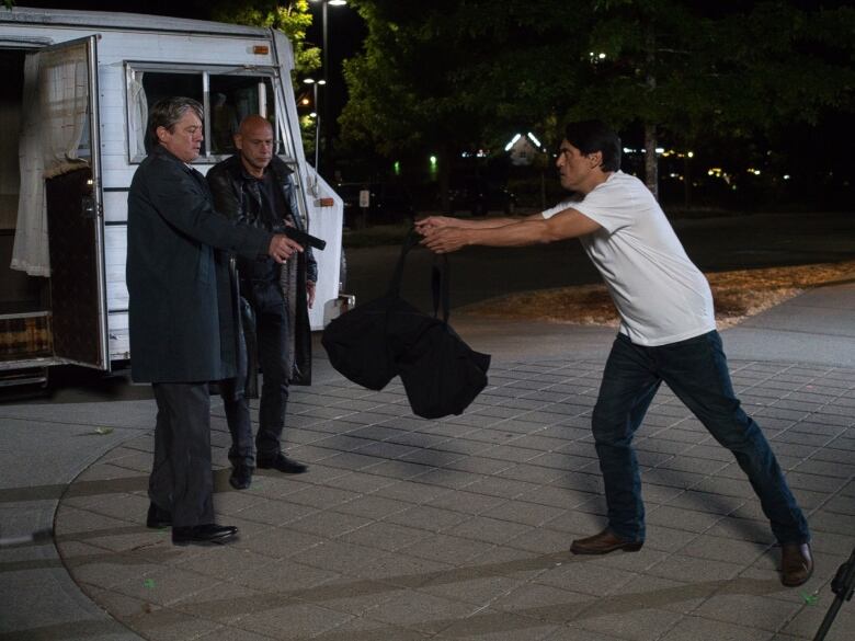 A man points a gun at one man while the other holds out a big black bag