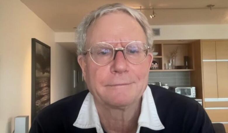 Dr. James Talbot is wearing glasses, a black sweater and white collard shirt as he looks directly into the camera. A kitchen can be seen behind him