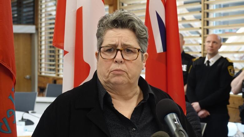 A woman with short grey hair stands at a microphone with a somber look on her face -- a deep frown. Behind her are the flags for Ontario and Canada.