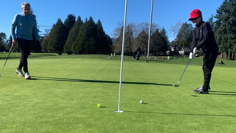 A woman hits a golf ball with her club on a putting green, toward a flag in a hole.