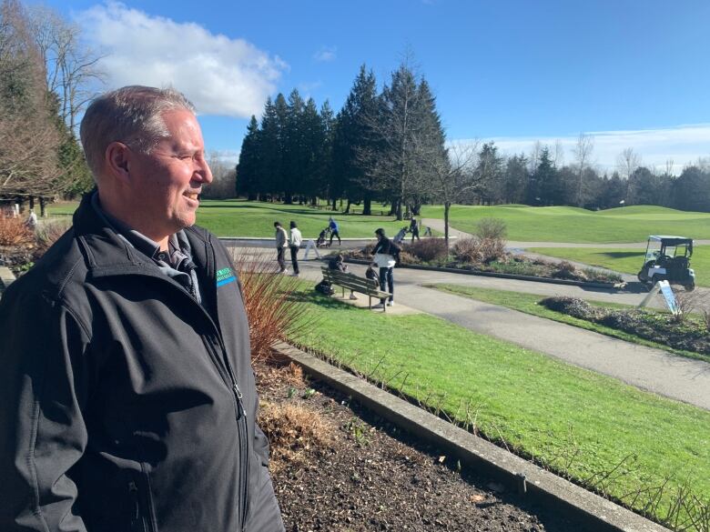 A man in a Vancouver golf course jacket looks out over a golf course with a golf cart and several golfers visible.