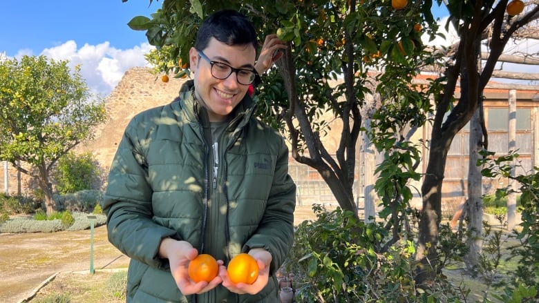 A young man in a warm coat smiles while holding oranges while standing in front of the orange tree that he picked them from. 