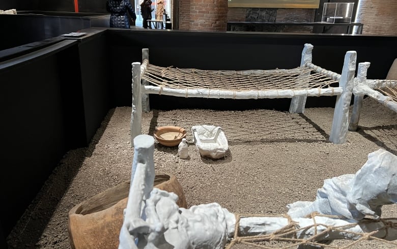 A museum exhibit shows bed frames with a rope net for lying on, along with pottery and other artifacts scattered on a pebbled floor.