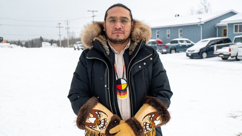 A person wearing a dark winter jacket and moose-hide gloves with a fur trim and beaded eagles on the front of them stands outside on a snowy day.