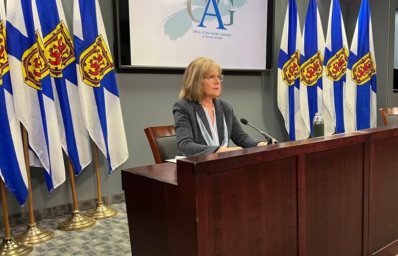 A woman with glasses sits at a desk with Nova Scotia flags behind.