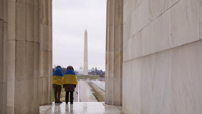 A couple draped in Ukrainian flags overlooking the Washington Monument. Image shows them from behind, framed inside the marble columns of the Lincoln Memorial, with the Washington obelisk in the background.