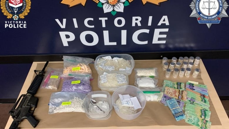 A table in front of a Victoria Police logo shows a gun, multiple containers filled with drugs and a stack of paper money.