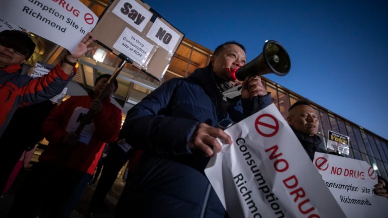 A man holds up a megaphone to his mouth, with people around him holding signs reading 'No Drug Consumption Site in Richmond'.