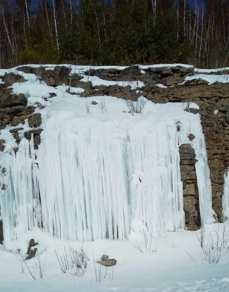 White streams of water are frozen against a rocky wall. The ground is covered in snow.