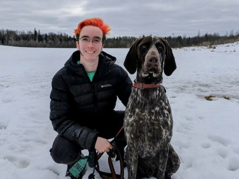 A person with orange dyed hair, wearing a black puffer jacket, kneels close to a brown and white dog.  