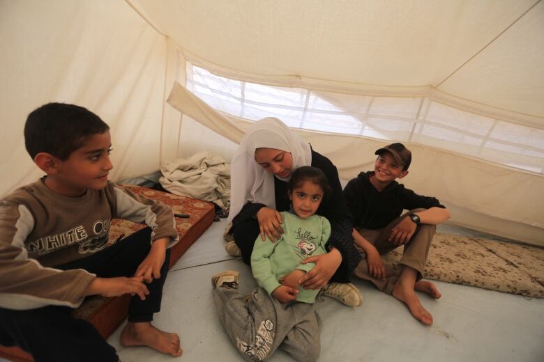 A young woman wearing a white hijab sits in a tent with her younger sister in her lap as her two younger brothers look on. 