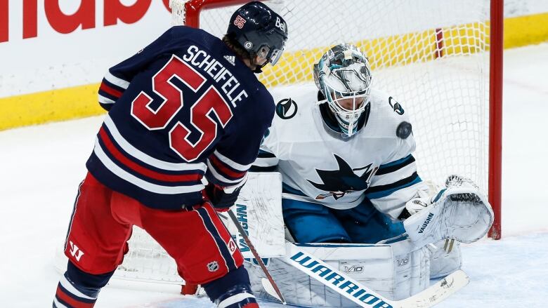 A hockey player in a blue and red uniform shoots a puck at a goalie in a white and teal uniform.