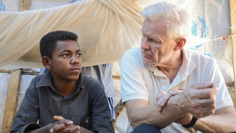 An old man sits in a makeshift tent with a young man.