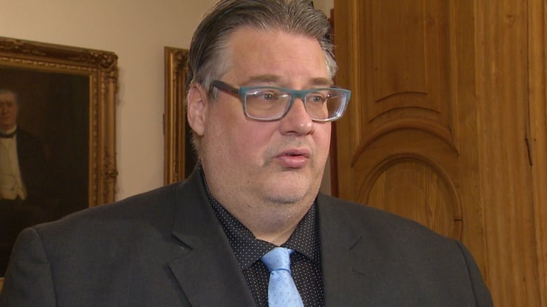 A man, seen from the shoulders up, with glasses wearing a black suit with a baby blue tie.