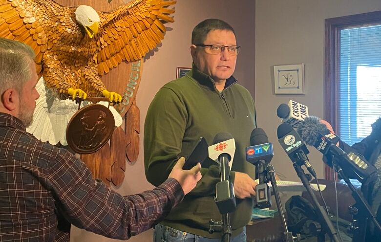 A man wearing a green sweater can be seen standing with reporters all around him holding mikes and asking questions. 