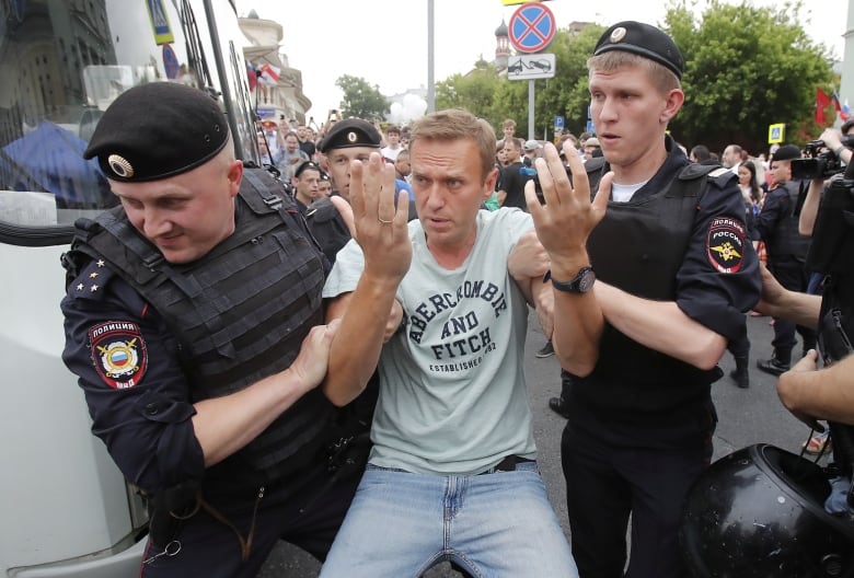 A man in a tshirt is taken away by police at what appears to be an outdoor demonstration.