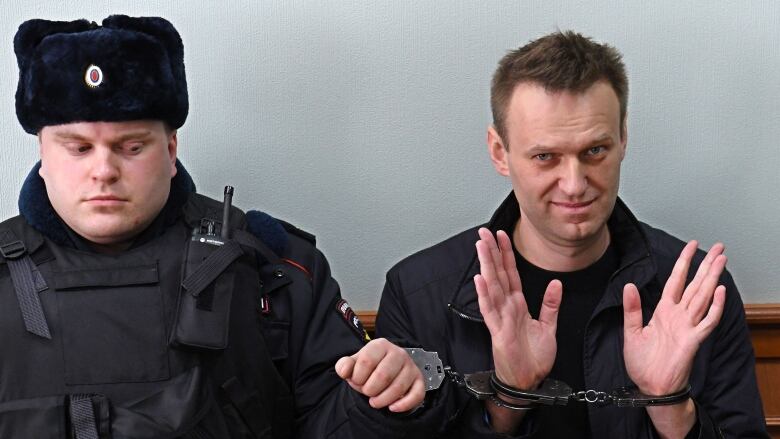 A man with a rye look on his face holds up his open, handcuffed hands. He is cuffed to a man in a uniform wearing an ushanka.