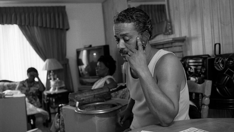 A black-and-white photo of a man holding an old cellphone to his ear. He's sitting at a kitchen table. Two women sit in the living room behind him.