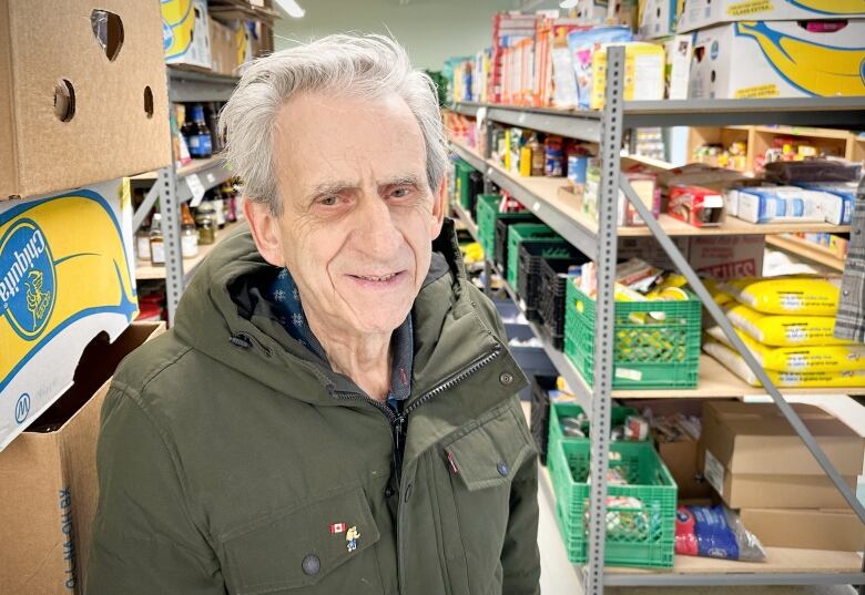 A man with grey hair wearing a green parka smiles and stands among shelves stacked with food.