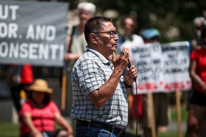 A man speaks into a microphone with demonstrators holding signs behind him.