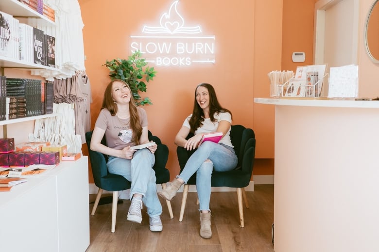 Two women sit in armchairs laughing and holding books. Above them is a neon sign with the words 