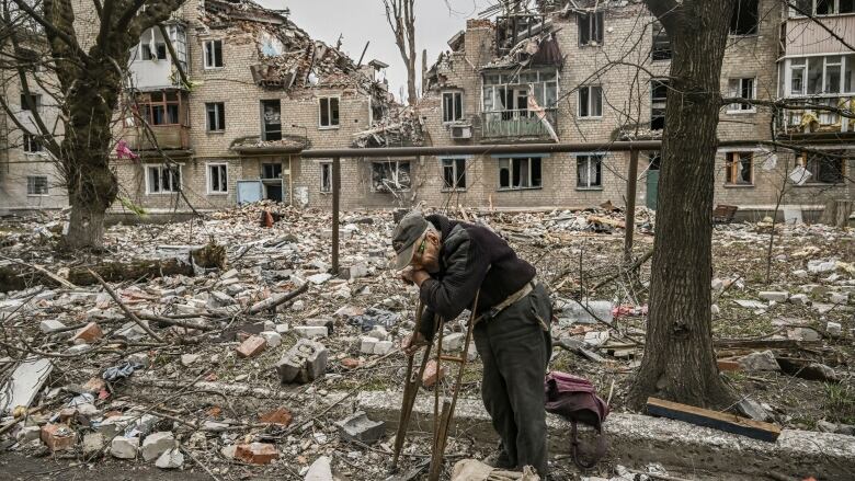 A person in crutches wipes their face with their hand as they stand amid debris and near a building destroyed by bombardment.