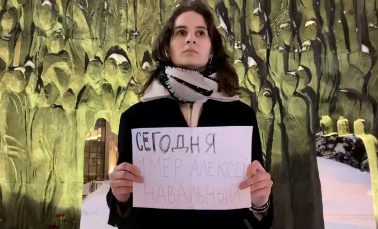 A woman with dark hair wears a winter jacket as she holds a white piece of paper with Russian writing on it in front of a memorial in Moscow. 