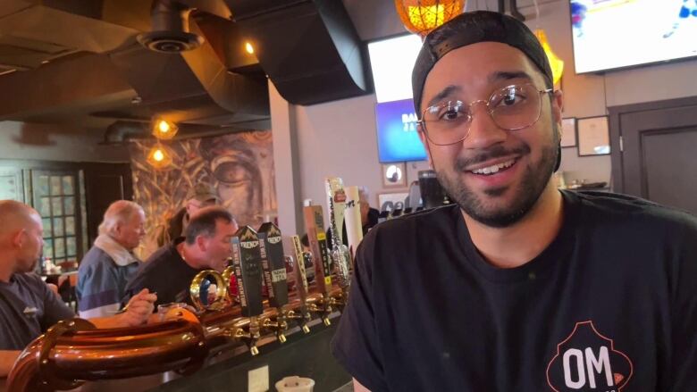 A man behind a bar talks to the camera while a group of men eat behind him.