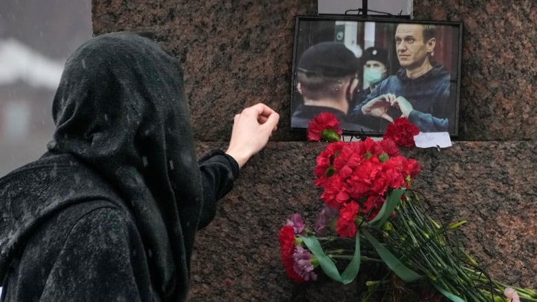 A person dressed in black touches a photo of a man above a bouquet of red flowers.