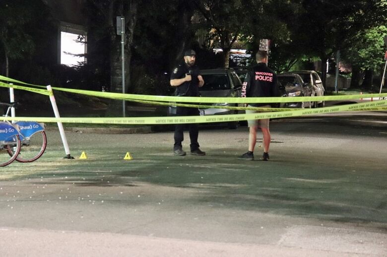 Two men in police uniforms stand behind a crime line, with yellow markers on the ground.