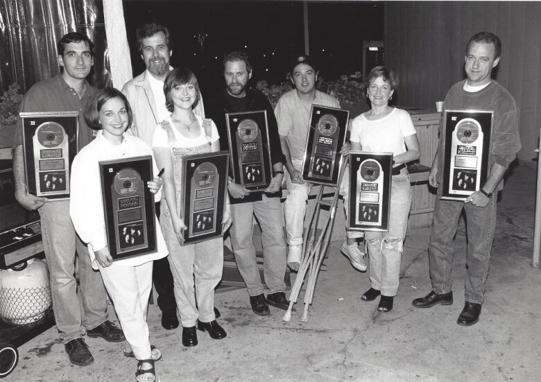 A black and white photo shows eight people, six of whom are holding plaques celebrating large record sales of an album.