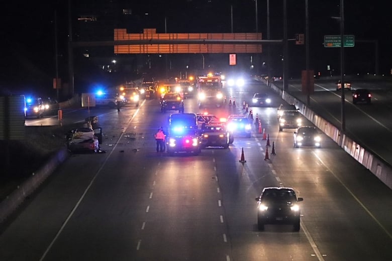 Emergency cars flash their lights around a pair of crashed vehicles located on the shoulder of a highway.