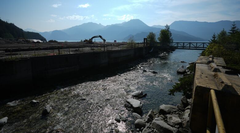 A scoop shovel works on a patch of dirt next to a picturesque river and mountains, with a river in the foreground.