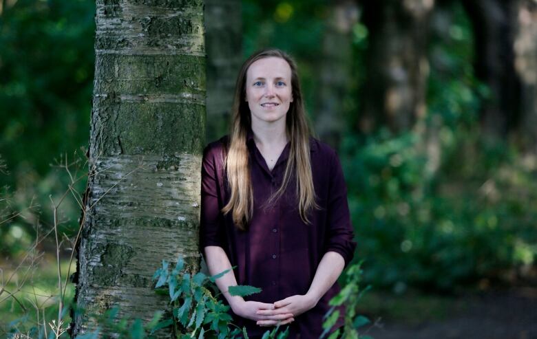 A smiling woman with long blond hair stands in a forested area leaning against a tree. 