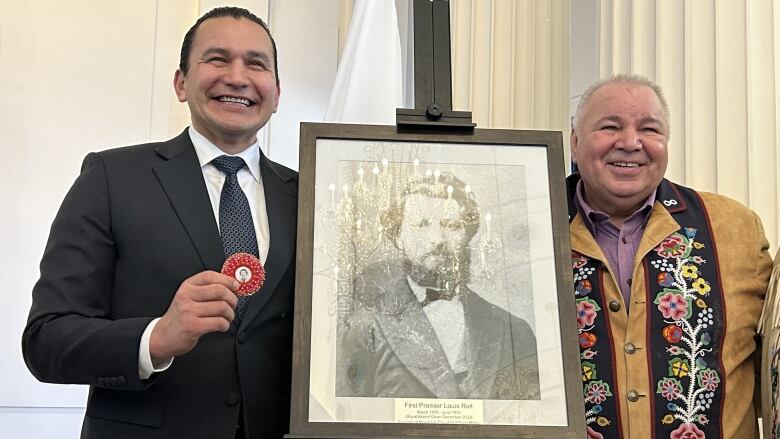 Two men stand side by side next to a framed portrait.