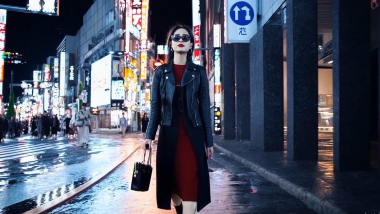 A woman walks on a Tokyo street at night with neon signs behind her. She wears a black leather jacket and red lipstick.