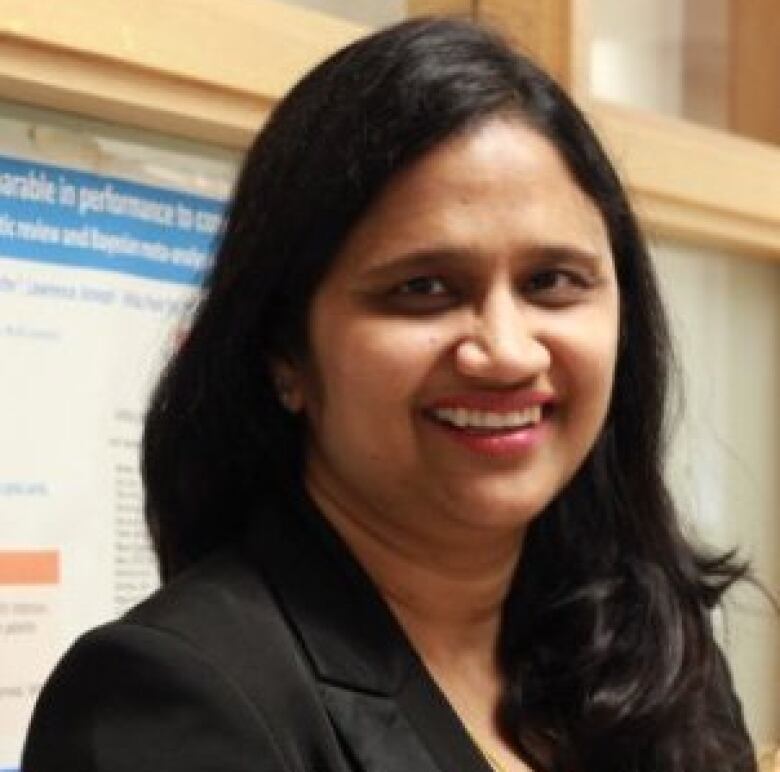 Dr. Pant Pai is wearing a black blazer and standing in front of a scientific poster