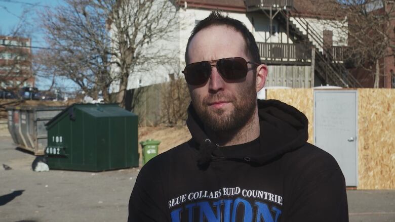 A man in sunglasses stands in front of a plywood structure. 