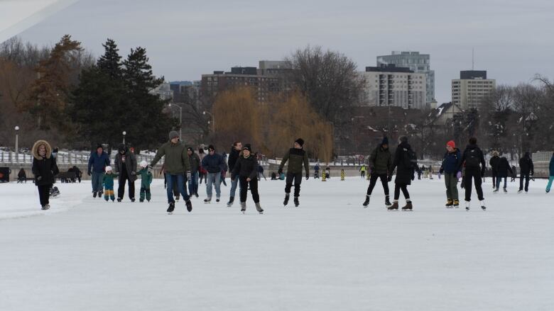 A row of people walk and skate along a large section of ice. Behind them are trees and large buildings.