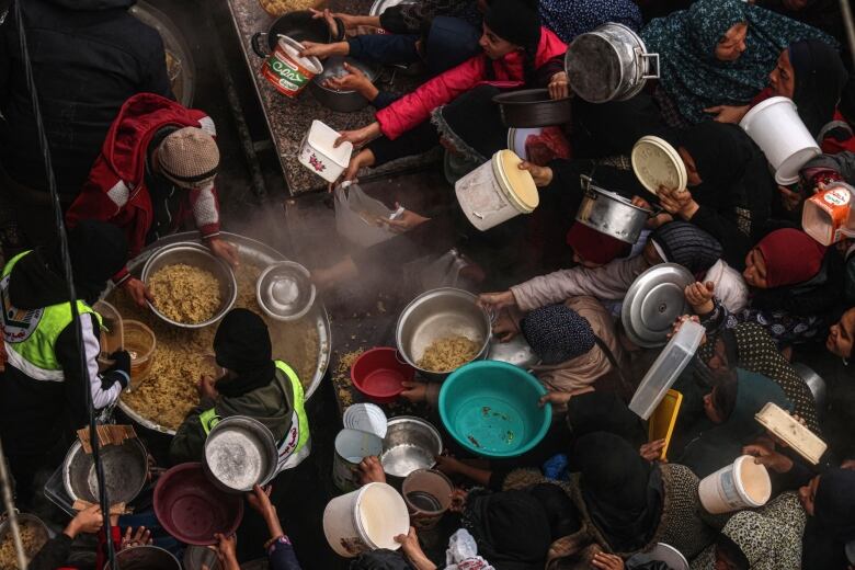 Women holding plates, bowls and buckets crowd together in front of people distributing food from a large metal pot. 