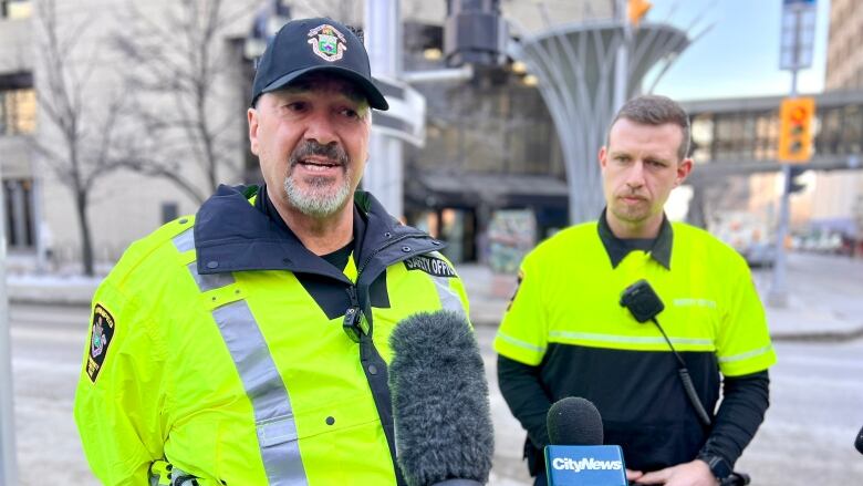 A man wearing a bright yellow jacket with a patch on the arm is standing on a sidewalk and speaking. Another man with a yellow shirt is standing next.