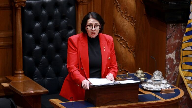 A white woman with black hair reads from an ornate seat in a legislature building.