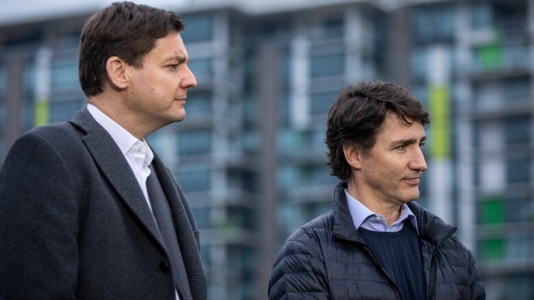 Two tall white men stand next to each other, with a housing complex in the background.