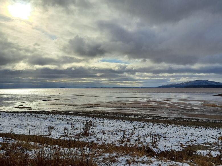 A silvery expanse of water beneath a cloudy sky. There is shadowy hill on the horizon.