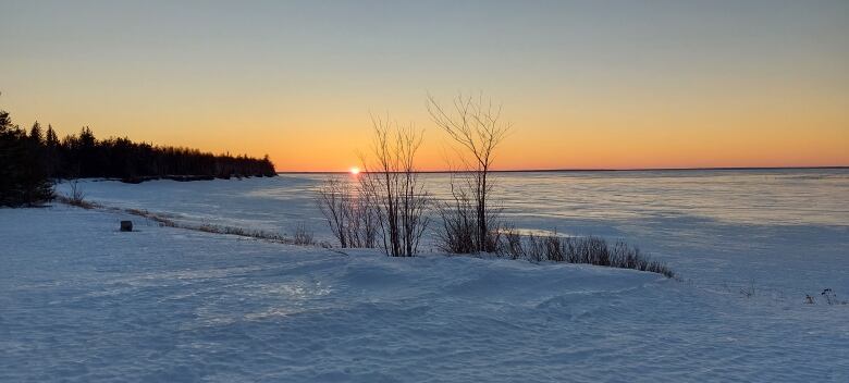 A sun slips down under the horizon over a snowy bay, sending orange colours up into the sky.