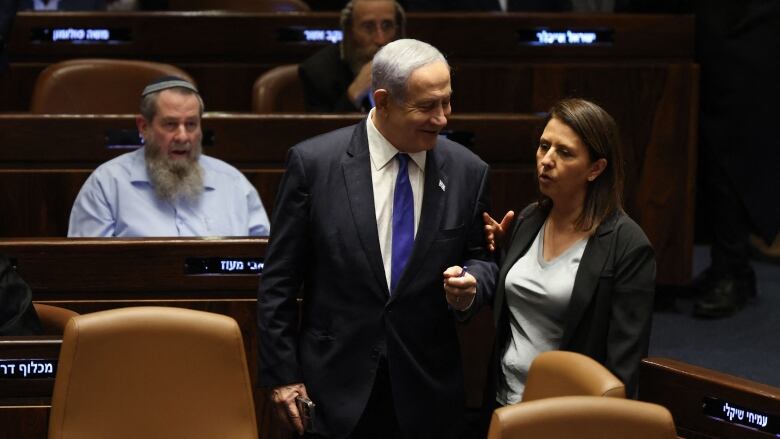 A cleanshaven older man in a suit and tie stands with left fist clenched in what appears to be a legislative chamber, with men and women shown seated behind him.