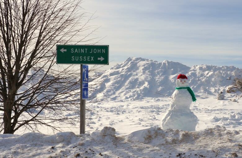 A snowman wearing a green scarf and a red hat stands next to a road sign that says Saint John and Sussex on it. There are massive snow piles behind him. 