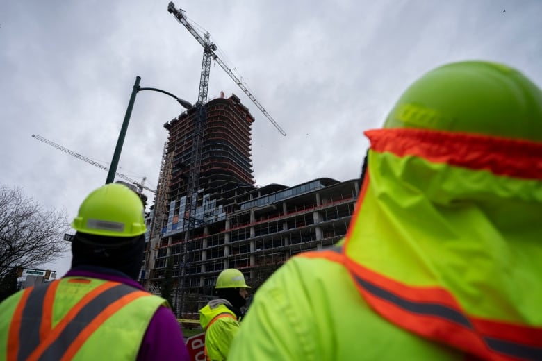 People in safety vests stare up at an under-construction building with a tower crane next to it.